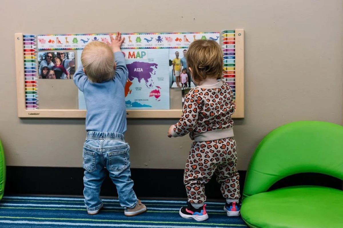 Two babies standing at a wall-mounted activity board